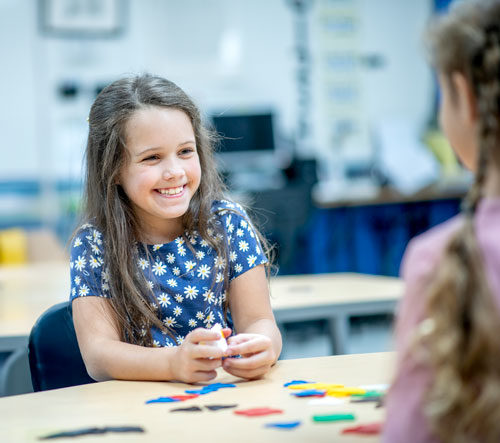 Children sitting at table across from eachother talking.