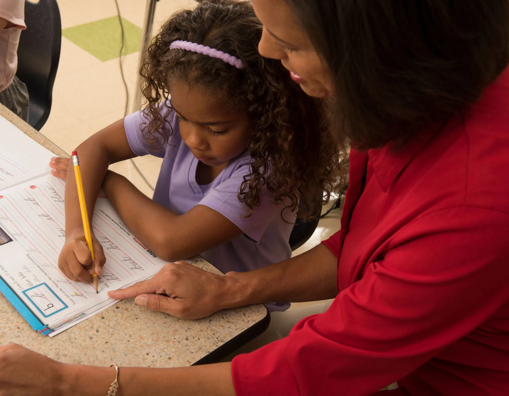 Child writing at a desk with teacher watching