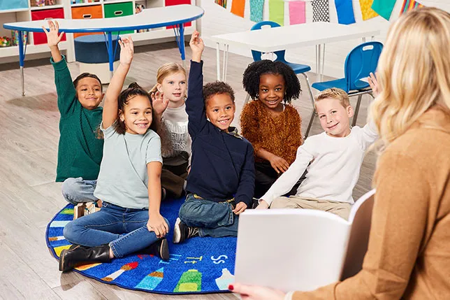 Small group of students, some with hands raised, on a rug facing teacher who is holding a book