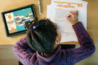 Student completing vocabulary page of Spelling Connections lesson with a spelling quest visible on nearby tablet