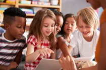 Students listening to whole-class read-aloud. One points to a book page and speaks while teacher is smiling.